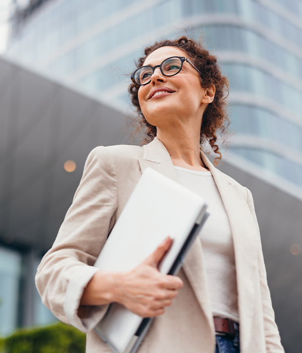 Smiling businesswoman holding laptop and looking away near office building